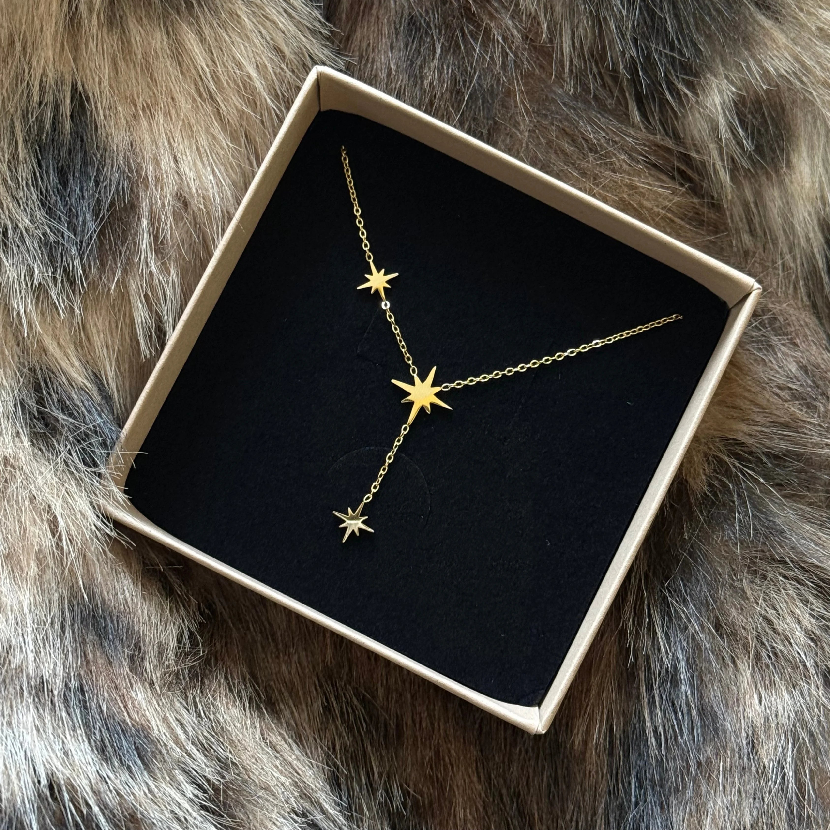 Collier fin forme Y MAISON DE L'AMOUR orné d'étoiles dorées, présenté dans son écrin cadeau sur fond texturé.