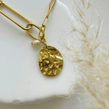 Gros plan sur la médaille texturée effet martelé et la breloque perle du collier posé sur un décor naturel.