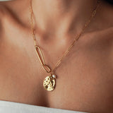 Collier chaîne maille trombone en acier inoxydable doré avec médaille martelée irrégulière et petite perle blanche, porté au cou.