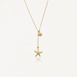 Collier lariat acier inoxydable doré avec pendentifs coquillage et étoile de mer, sur fond blanc.