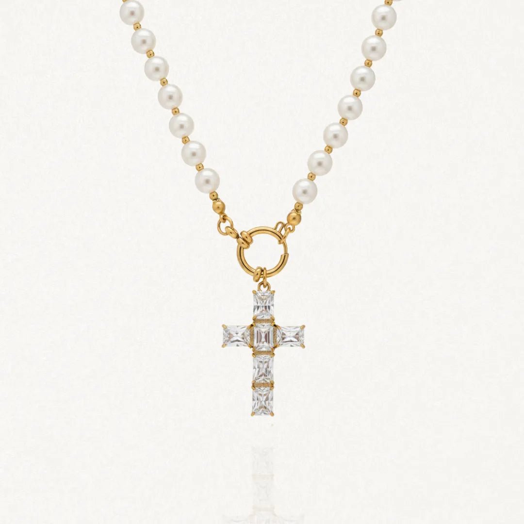 Collier doré en acier inoxydable avec une chaîne composée de perles rondes blanches, reliée par un fermoir toggle. Un pendentif en forme de croix est suspendu, entièrement pavé de zircons rectangulaires brillants.