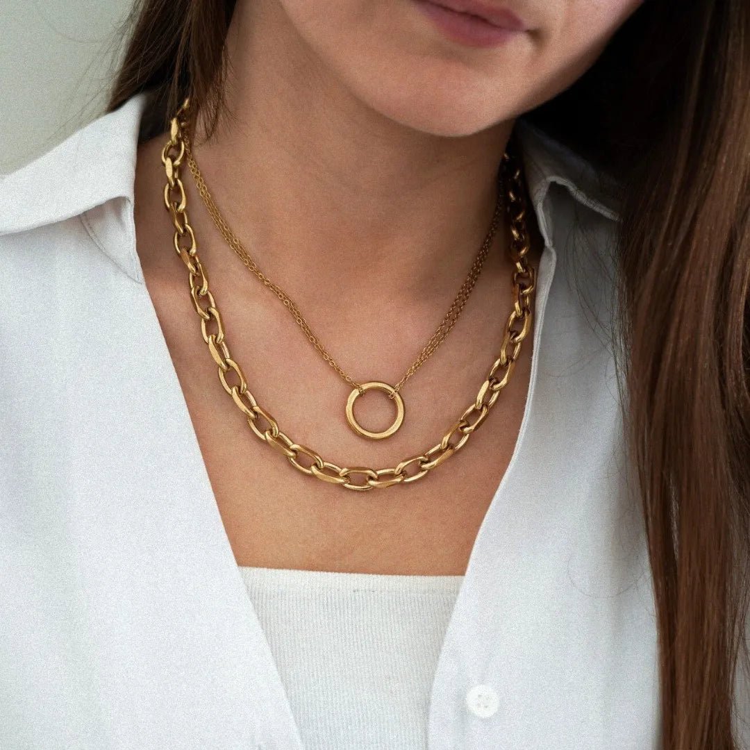 Femme portant deux colliers en or superposés: une chaîne épaisse à maillons et un collier fin avec pendentif anneau, sur une chemise blanche.