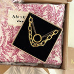 Collier doré double chaîne (grosse à maillons ovales, fine avec anneau) dans écrin noir sur tissu motif floral rose, étiquette AMOU.