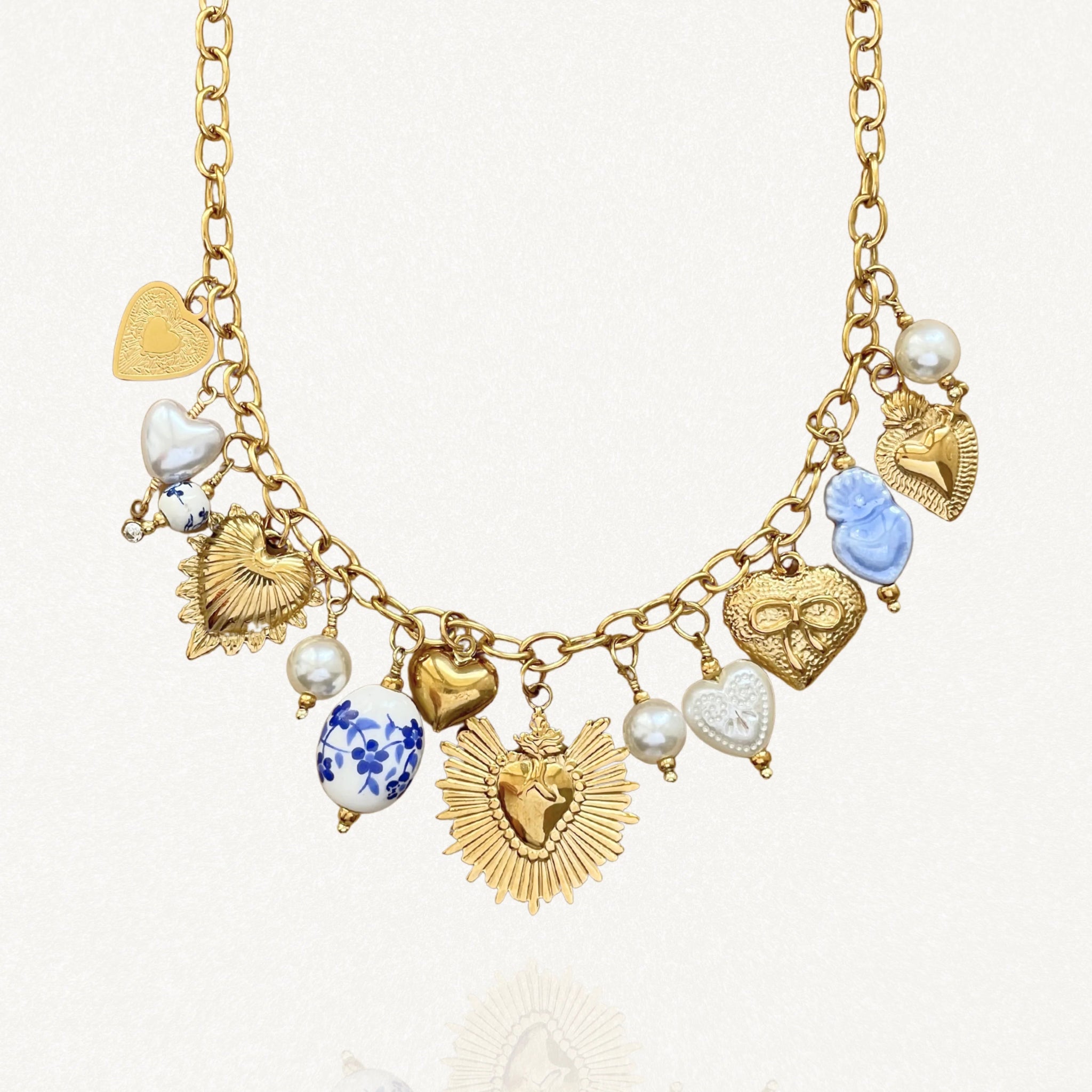 Gros plan du collier chaîne en acier inoxydable doré, montrant en détail les breloques : cœur sacré ex-voto avec œil, perles en céramique bleue et perles nacrées. Bijou résistant à l'eau.