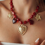 Collier statement à grosses perles rouges et multiples breloques dorées (cœur sacré, soleil, étoile, rose) porté au cou.