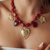 Collier statement à grosses perles rouges et multiples breloques dorées (cœur sacré, soleil, étoile, rose) porté au cou.