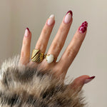 Duo de bagues dorées portées à l'index et au majeur, mises en valeur par une manucure graphique rouge.