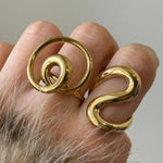 Accumulation de deux grosses bagues dorées sculpturales (modèle spirale et modèle vague) portées sur la même main.