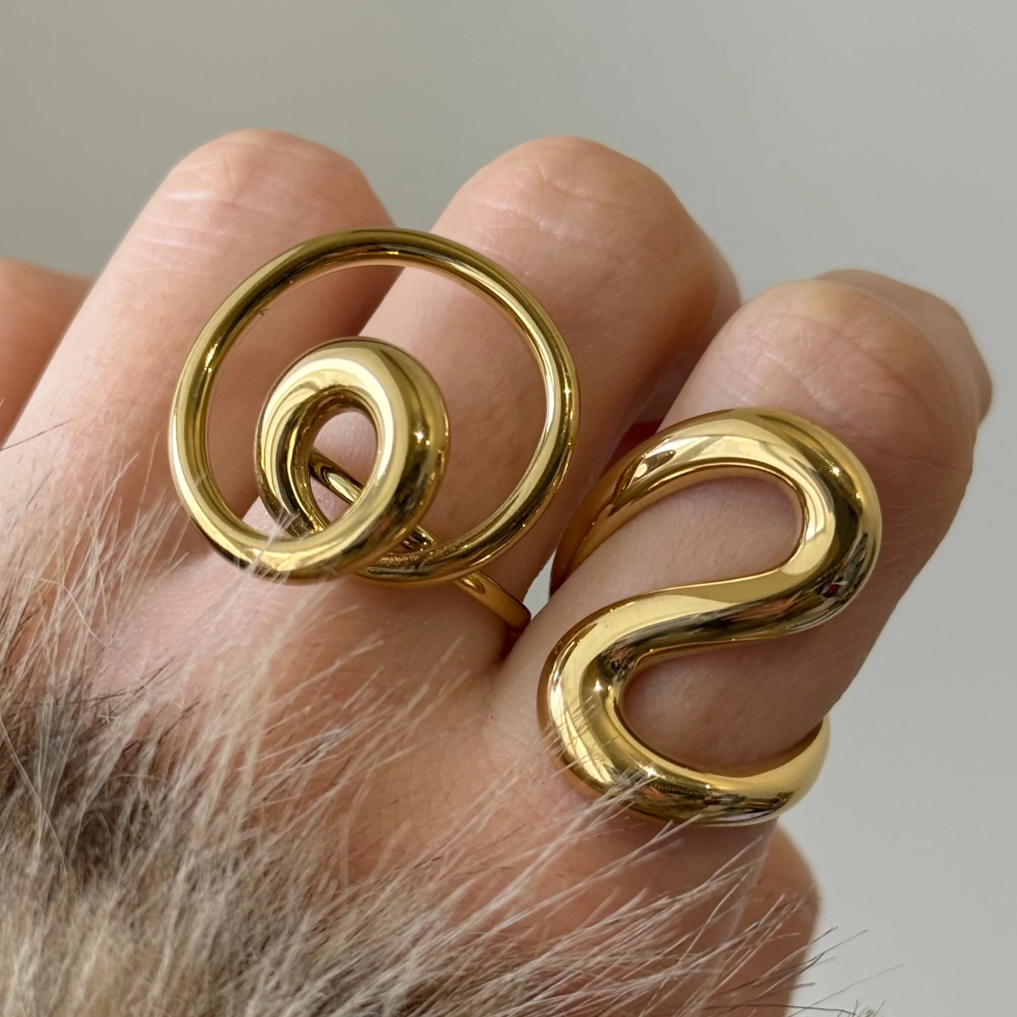Accumulation de deux grosses bagues dorées sculpturales (modèle spirale et modèle vague) portées sur la même main.