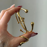 Main tenant le bracelet jonc ouvert et portant une bague dorée assortie au design organique et fluide.