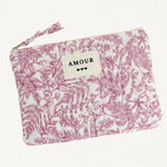 Pochette rectangulaire en tissu matelassé blanc à motif floral rose foncé. Une étiquette beige cousue au centre porte l'inscription 'AMOUR' et trois petits cœurs noirs. Fermeture éclair avec tirette en tissu.