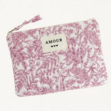 Pochette rectangulaire en tissu matelassé blanc à motif floral rose foncé. Une étiquette beige cousue au centre porte l'inscription 'AMOUR' et trois petits cœurs noirs. Fermeture éclair avec tirette en tissu.