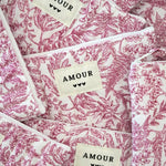 Multiples trousses matelassées en tissu blanc imprimé toile de Jouy rose fuchsia, étiquettes crème 'AMOUR' et 3 cœurs noirs.