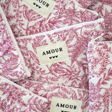Multiples trousses matelassées en tissu blanc imprimé toile de Jouy rose fuchsia, étiquettes crème 'AMOUR' et 3 cœurs noirs.
