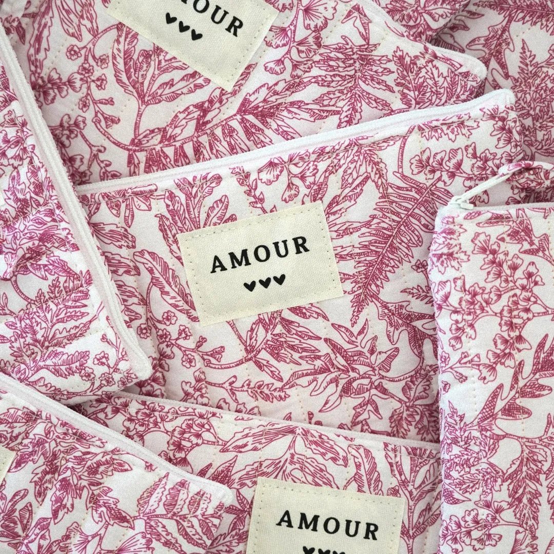 Multiples trousses matelassées en tissu blanc imprimé toile de Jouy rose fuchsia, étiquettes crème 'AMOUR' et 3 cœurs noirs.