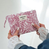 Mains féminines aux ongles blancs avec bagues dorées, tenant une pochette matelassée blanche à motif floral rose/rouge AMOUR.