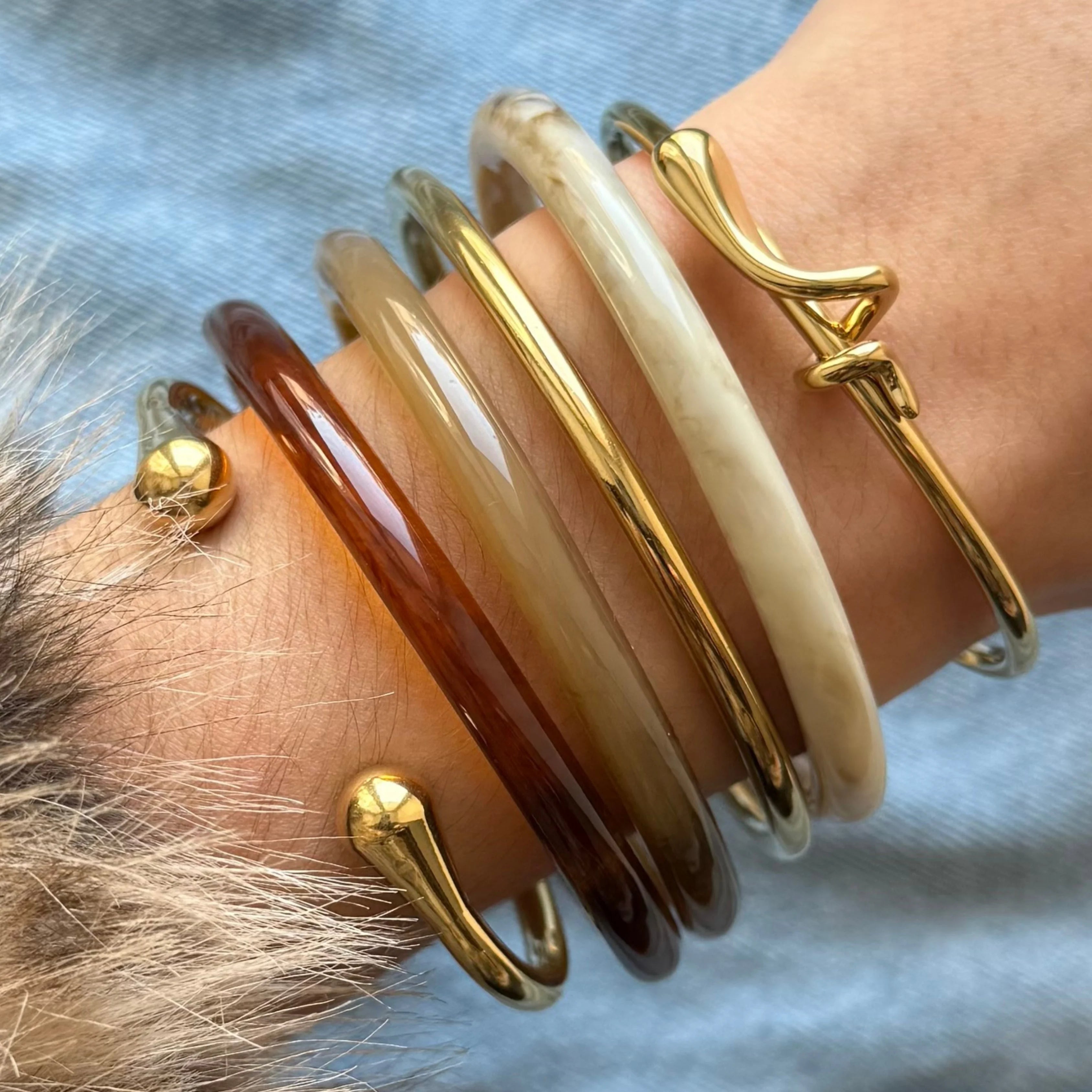 Idée de stacking bijoux : accumulation de joncs en résine mélangés avec des bracelets joncs ouverts dorés (modèles nœud et boules) au poignet.